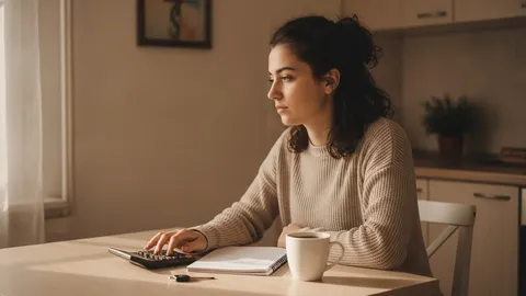 Tânără într-un pulover bej stă la masa din bucătărie, lângă fereastră, și calculează pe un calculator de birou, alături de un carnețel deschis, o cană de cafea și o cheie de mașină.