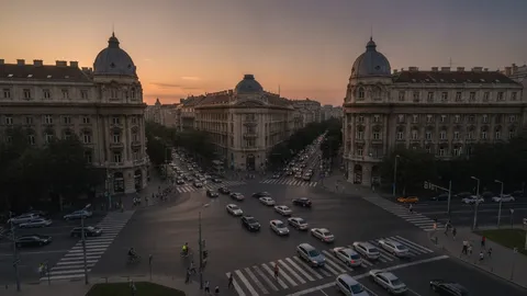 Intersecție aglomerată dintr-un oraș, văzută de sus, în lumina caldă a apusului, cu mașini oprite la semafor, pietoni care traversează pe trecere și un biciclist în mijlocul intersecției.