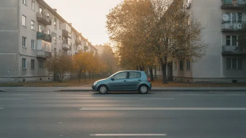 Hatchback compact albastru parcat lateral pe o stradă liniștită, cu blocuri comuniste și copaci cu frunze aurii de toamnă în fundal, într-o dimineață cu lumină caldă.