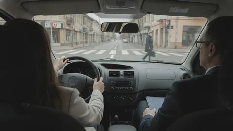 Vedere din spatele mașinii: candidata ține volanul cu ambele mâini, examinatorul stă pe scaunul din dreapta cu un dosar în poală, iar prin parbriz se vede o femeie pregătindu-se să traverseze pe trecerea de pietoni.