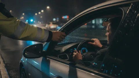 Polițist cu vestă reflectorizantă întinde etilotestul prin geamul lăsat al mașinii, către un șofer cu mâinile pe volan, oprit noaptea în trafic sub lumina albastră a girofarului.