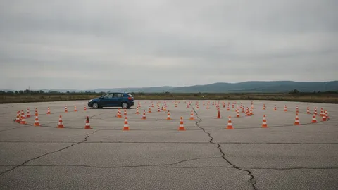 Poligon de școală de șoferi cu jaloane portocalii pe asfalt crăpat și o mașină mică albastră, văzută din lateral, în depărtare, sub un cer înnorat.