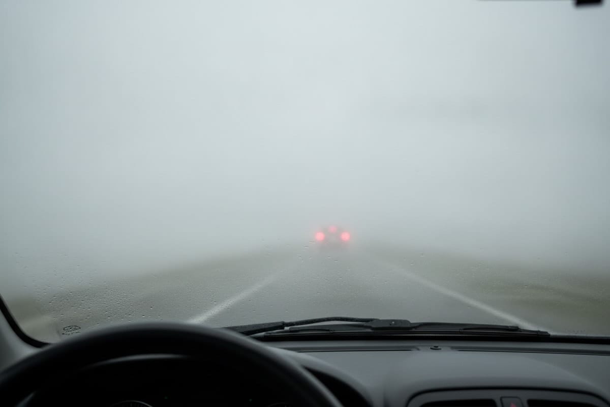 Vedere din interiorul mașinii, prin parbrizul cu picături de ploaie, pe ceață densă, cu stopurile mașinii din față abia vizibile și ștergătoarele în repaus la baza parbrizului.